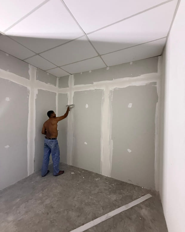 A worker skim-coating jointed plasterboard partitions with a float, under a newly installed suspended ceiling grid.