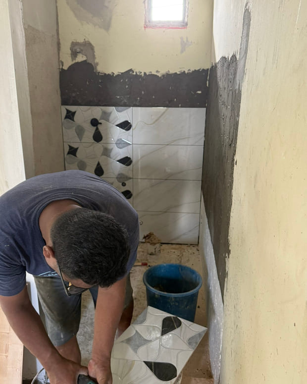A tiler crouched over a bucket, buttering a black-and-white leaf-pattern feature tile, with the half-tiled wall behind.