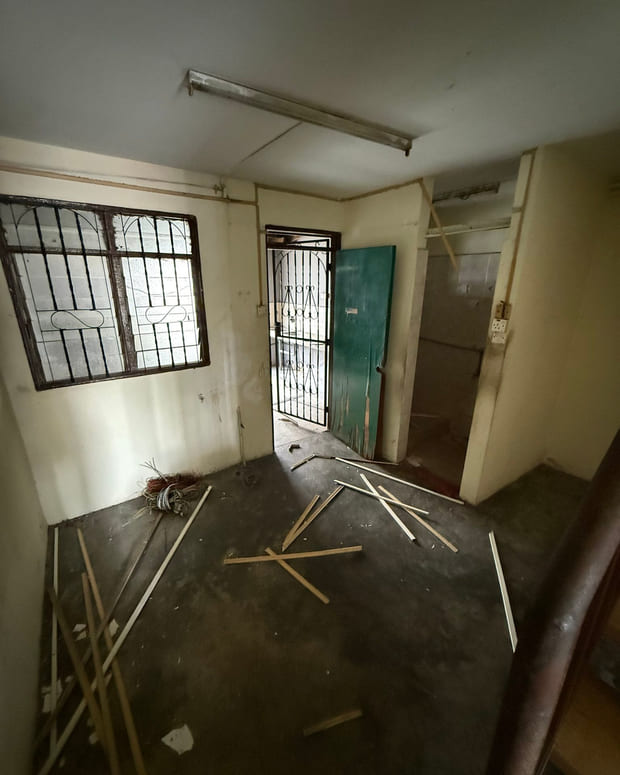 A bare room before work: a cracked cement floor, grilled windows, an old green-painted timber door and flaking cream paint.