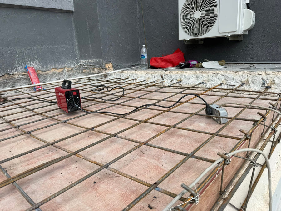 A steel reinforcement mesh tied over an old tiled floor, threaded through with grey electrical conduit and a white waste pipe.
