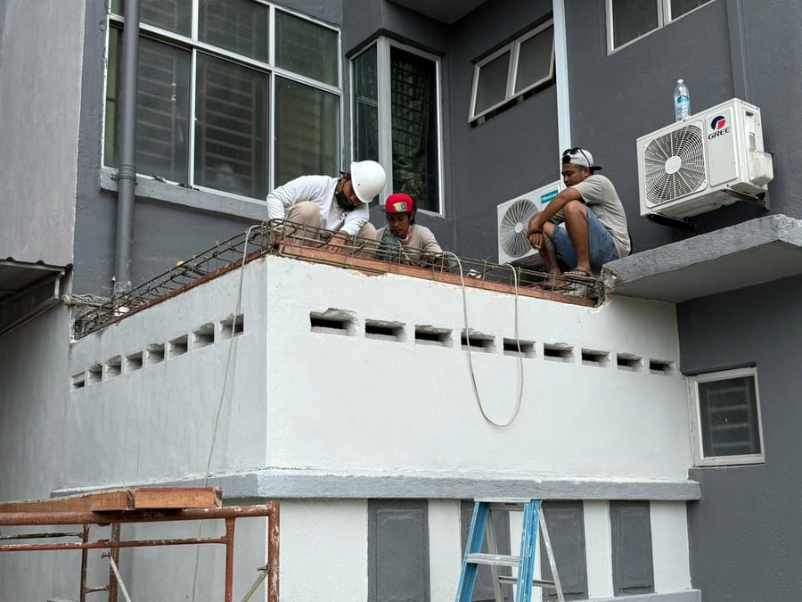 Three workers on top of a newly built, rendered ground-floor extension against an apartment block, tying steel reinforcement along its walls.