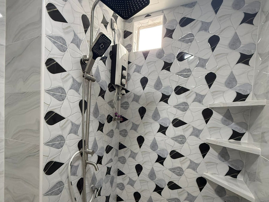 A full-height shower wall in black-and-grey leaf-pattern tile with three stepped corner shelves formed into it, a black rain-shower set and an instant water heater.